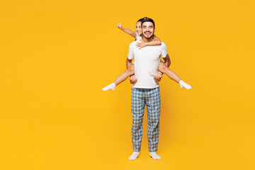 Full body young cool fun parent man with child girl wear blue pyjamas clothes. Daddy little kid giving piggyback ride to joyful isolated on plain yellow background. Father's Day Love family concept.