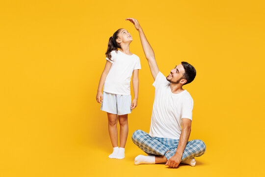 Full body young parent man with child girl wear casual blue clothes. Daddy little kid hold hand above head show how much she grown isolated on plain yellow background Father's Day Love family concept