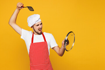 Young housewife housekeeper chef cook baker man wear red apron toque hat hold in hand spatula lid pov fight with shield isolated on plain yellow orange background studio portrait Cooking food concept