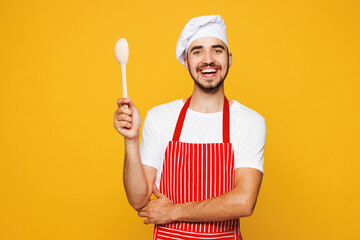 Young smiling happy fun housewife housekeeper chef cook baker man wear red apron toque hat hold in hand wooden spoon isolated on plain yellow orange background studio portrait. Cooking food concept.