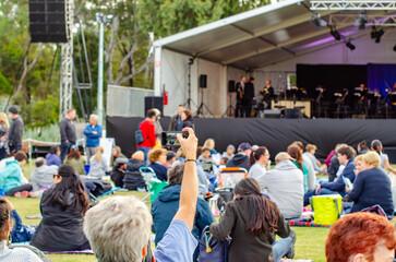 A raised hand holding a phone captures the crowd gathered at an outdoor live music event in a public park, people sitting in front of a performance stage. A lively community atmosphere in Australia.