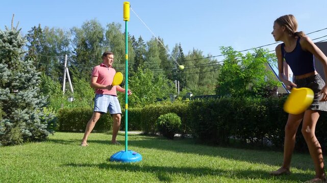 A father and daughter enjoy a sunny day playing swingball (tetherball) in their garden, laughing and having fun