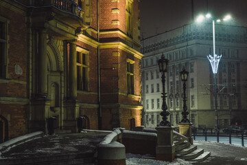 View of an old red brick building with ornate details and snow-covered steps under the glow of streetlights at night, Gdansk, Poland.