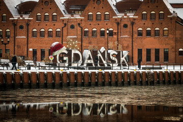 View of the iconic 'GdaÅ„sk' sign adorned with a Santa hat, nestled against the backdrop of brick buildings reflecting in the MotÅ‚awa River, GdaÅ„sk, Poland.