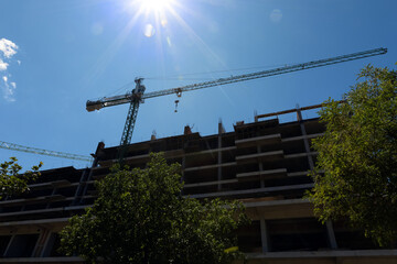 Silhouetted view of a building under construction with a green tower crane, captured under bright sunlight and a clear blue sky.