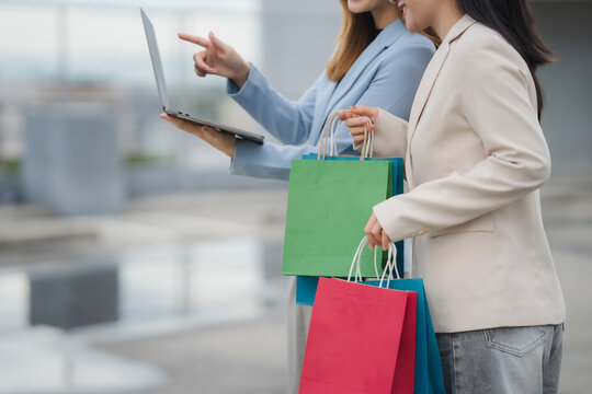 Window Shopping & Business: Two fashion-forward women, adorned in chic attire, collaborate over a sleek laptop while cradling vibrant shopping bags, perfectly capturing a fusion of commerce and style.