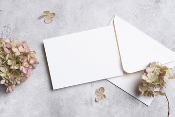 Blank wedding invitation card mockup with white envelope on textured gray background, surrounded by dried hydrangea flowers, copy space for card design