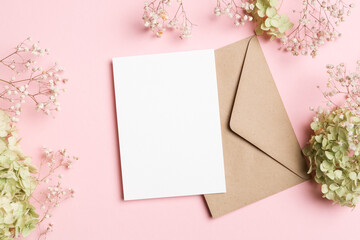 Blank greeting card mockup with brown envelope on pink background surrounded by delicate flowers, copy space for wedding card design