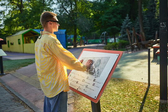 Blind man reading tactile information map in public park. Concept of assistive tech, urban accessibility, inclusive navigation, and social participation.