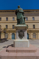 A weathered bronze statue of a historical figure in robes stands on a stone pedestal in a sunlit public square, with a light-colored building in the background.