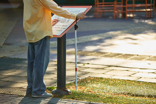 Blind man standing with cane and reading public tactile board in inclusive park. Concept of urban accessibility, social inclusion, assistive technology, tactile learning.