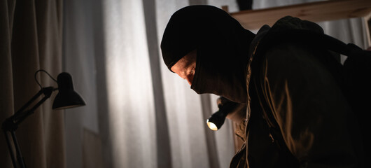 thief in black balaclava into apartment using flashlight searching money