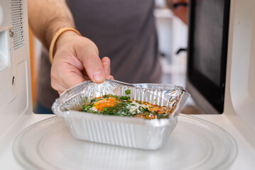 A person placing a foil takeout container with a meal (eggs and greens) into a microwave oven.