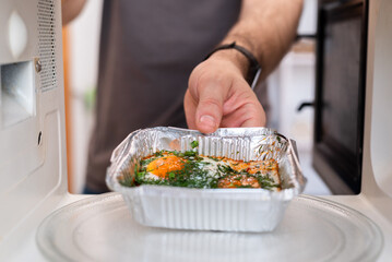 Close up of man placing foil food container with cooked dish into microwave oven.