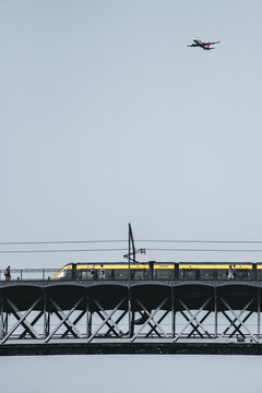 Porto, Portugal - 17 June 2022: View of a modern yellow train traversing the metallic Dom Lu&Atilde;&shy;s I Bridge as a plane flies above, a blend of transport modes against a muted sky.