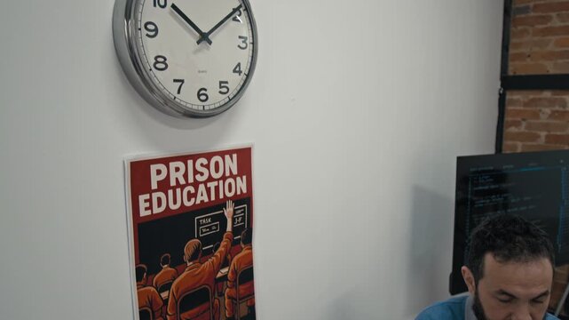 Medium shot of Caucasian male computer literacy teacher sitting at desk, working on laptop during lesson in prison classroom, under cheerful motivational poster and clock on wall