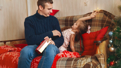 A little girl give surprise father with a gift box on the Christmas. Near the Christmas tree at home