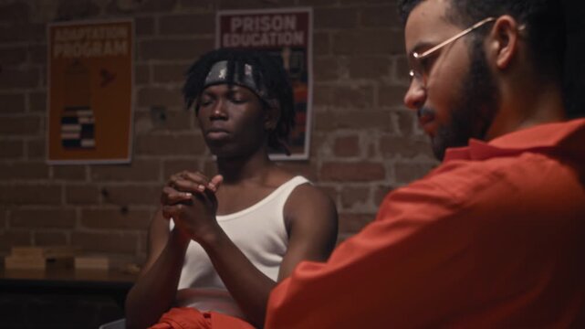 Medium shot of pensive African American and Middle Eastern male prisoners in orange jumpsuits sitting at therapy session, hands clasped, listening attentively to companions confessing to past crimes