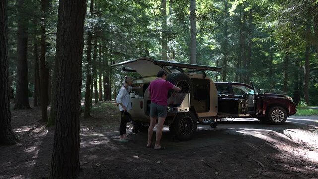 A man and woman are cooking breakfast together in a teardrop trailer at a serene campground, surrounded by towering trees under dappled sunlight.