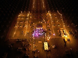Nighttime Gathering: A vibrant aerial view of an illuminated outdoor event, showcasing a multitude of tables, lights, and a central stage.