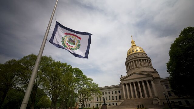 The West Virginia State Capitol showcases its impressive dome while the state flag waves gently. Surrounding trees add natural beauty to the historic site in Charleston.