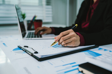 A businesswoman analyzes financial documents with charts and graphs at a desk, highlighting data trends with a pen in a bright, professional office setting.	