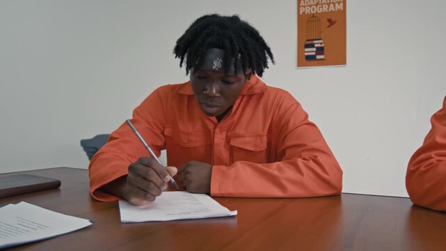 Medium pan shot of busy young Middle Eastern and African American male convicts in orange robes writing resumes, then sighing, while working hard during adaptation course for release on parole