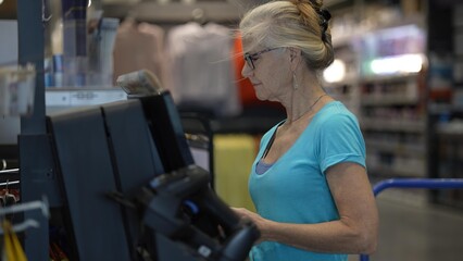A mature woman stands at a self checkout in a hardware store, scanning items and paying with a credit card while completing her shopping for home improvement supplies.