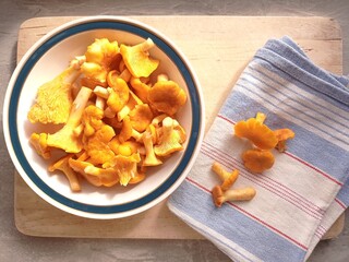 Top view of a porcelain bowl full of fresh and cleaned chanterelles. The bowl is on a wooden cutting board with a kitchen towel and a few more chanterelles.