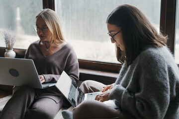 Two women are working on their laptops near a bright window. They are wearing comfortable clothes and working from their apartment on computers. The women appear focused on their screens.