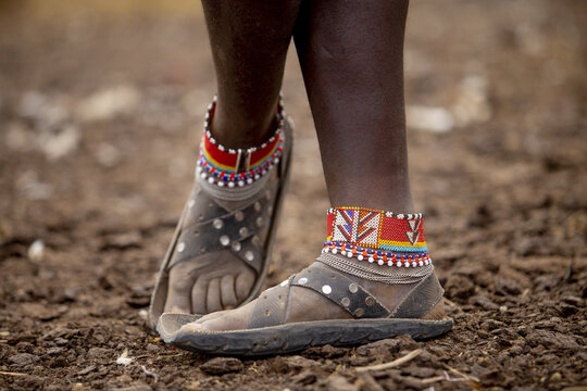 View of vibrant beaded anklets adorn dark skin above worn leather sandals, standing on the textured earthy ground, Narok, Narok, Kenya.