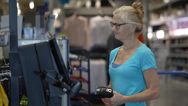 A confused, frustrated unhappy, mature woman stands at a self checkout station in a hardware store holding a hand scanner. - Powered by Adobe