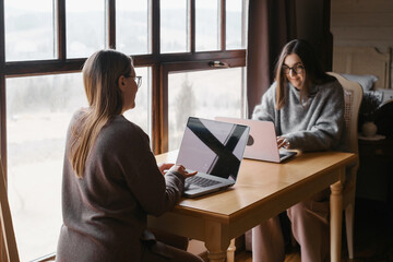 Two women working on laptops at a table near a large window. Comfortable and productive work environment, ideal for illustrating remote work, collaboration, or digital lifestyle concepts.