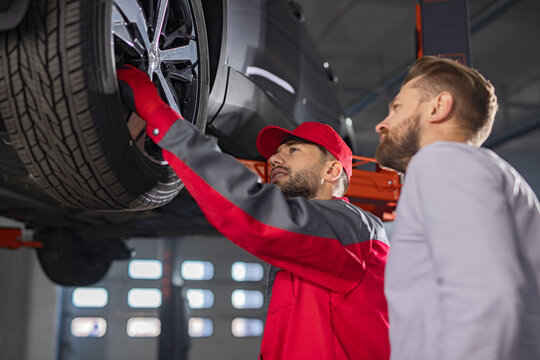 Mechanic carefully inspecting vehicle tires while guiding a customer in a professional auto repair shop. Scene emphasizing automotive expertise, maintenance, inspection, and customer service.