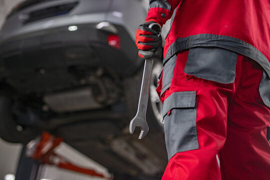 A mechanic wearing red and gray uniform holds a wrench beside a lifted vehicle in an automotive repair shop. The image represents vehicle maintenance, repairs, and professional automotive service