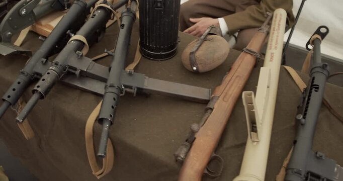 A collection of historic World War II era firearms and equipment on display. The table includes British Sten submachine guns, a German Panzerfaust anti-tank weapon, and other military artifacts. Conce