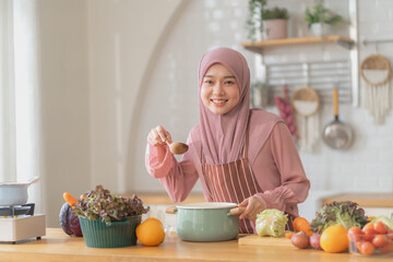 Smiling muslim woman in hijab and apron holding a spoon while cooking in a bright kitchen, surrounded by fresh vegetables, representing joy and warmth in muslim home cooking.