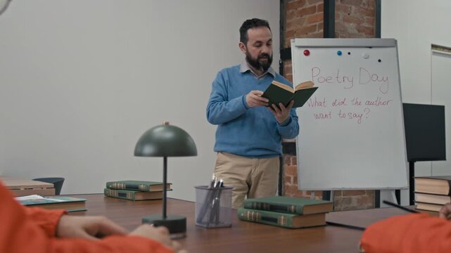Medium full shot of middle-aged Caucasian English literature male teacher conducting poetry lesson for group of young multiethnic prisoners in orange robes, reading verse from book