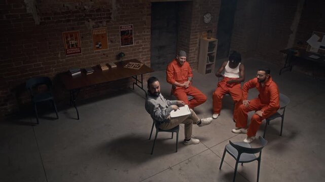 High-angle security camera shot of male prison psychologist and multiethnic convicts in orange uniforms arriving in recreation hall for group therapy session, sitting down on chairs in circle