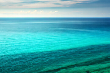Obraz premium Crystal clear turquoise ocean water stretching to the horizon under a soft blue sky with scattered clouds, reflecting the sunlight and creating a serene, tranquil natural beauty