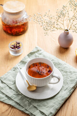 A cup of herbal tea with lemon and bouquet of flowers on a wooden table with teapot and napkin.
