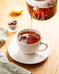 Herbal tea pouring from teapot into white cup on wooden table with lemon and napkin.