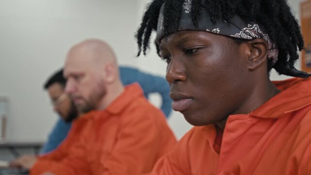 Tilting medium close-up of hands and face of busy young African American male convict in orange suit, with bandanna and braids typing on laptop while performing task at coding lesson in prison