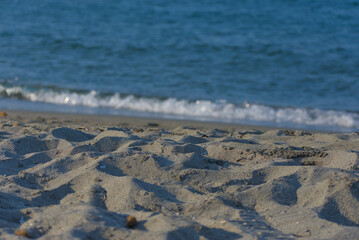 A close-up, low-angle shot of textured sand on a sunny beach with gentle, blurry waves of the sea visible in the background, creating a calm and natural mood.