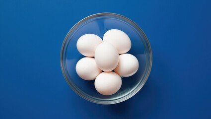 Overhead shot of fresh white eggs in a glass bowl on blue background