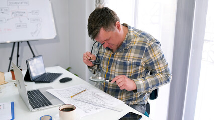 A middle-aged man examines a piece of paper with a drawing using a magnifying glass. The chief architect is working on a large construction project