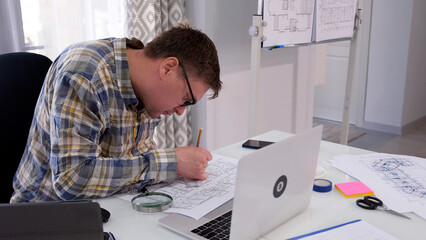 A stooped man with glasses is sitting at a table with a laptop and building drawings. The working days of a civil engineer. An architectural college student