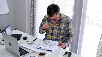 The engineer checks the building plans. A man in glasses and a plaid shirt holds a pencil in his mouth and carefully studies the plans of the structures