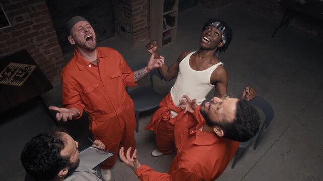 Medium top view of male prison counselor and multiethnic convicts in orange jumpsuits screaming together at therapy session to release negative emotions, then clapping with joy and relief