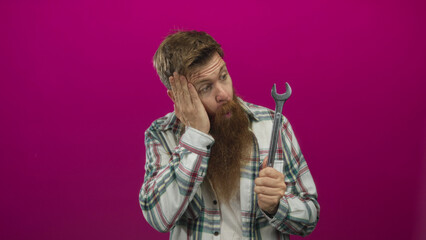 Man holds wrench with open mouth and raises hand to cheek in studio against magenta backdrop; surprise.